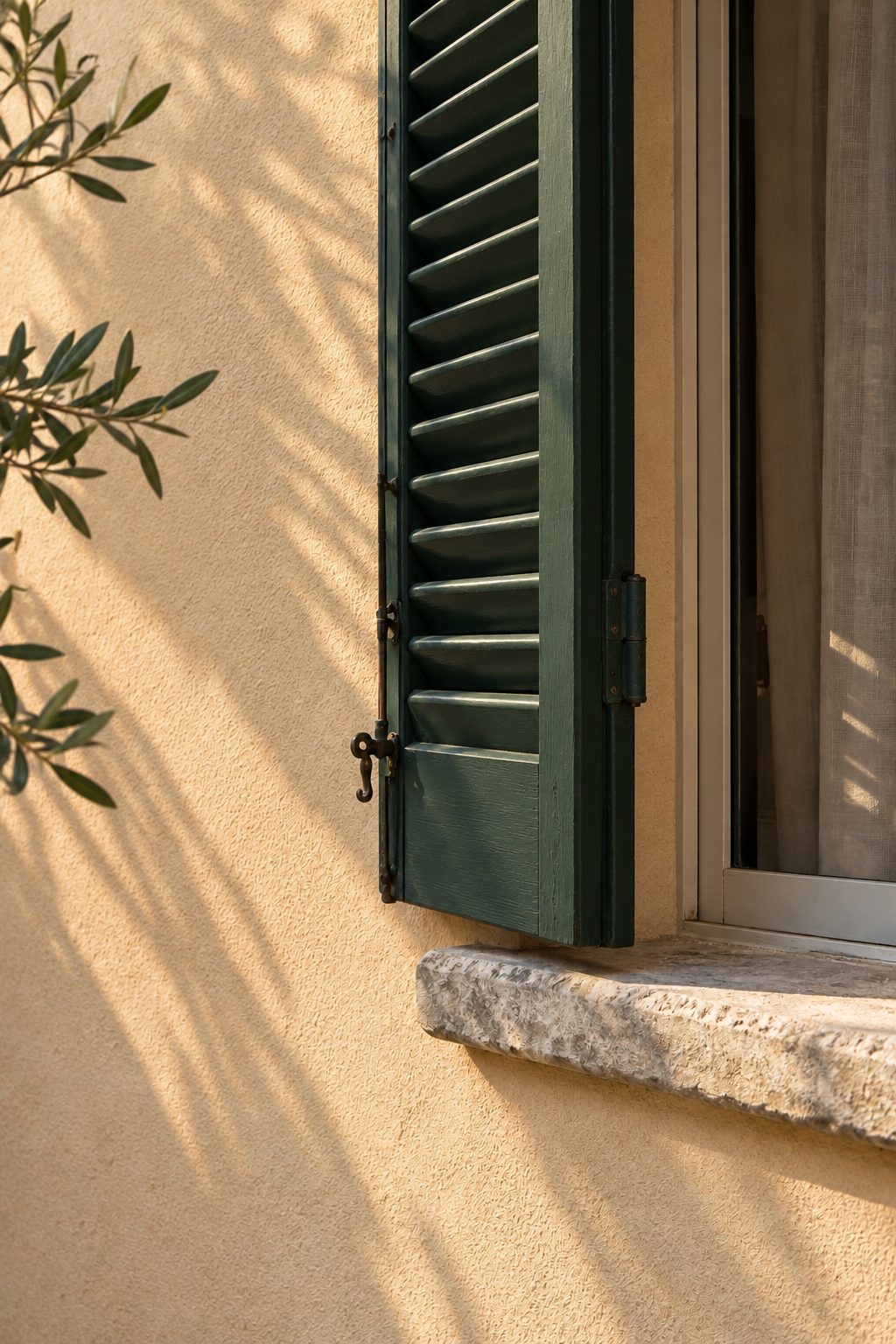 Finestra con persiana verde e davanzale in pietra su facciata in intonaco, con un ramo di olivo